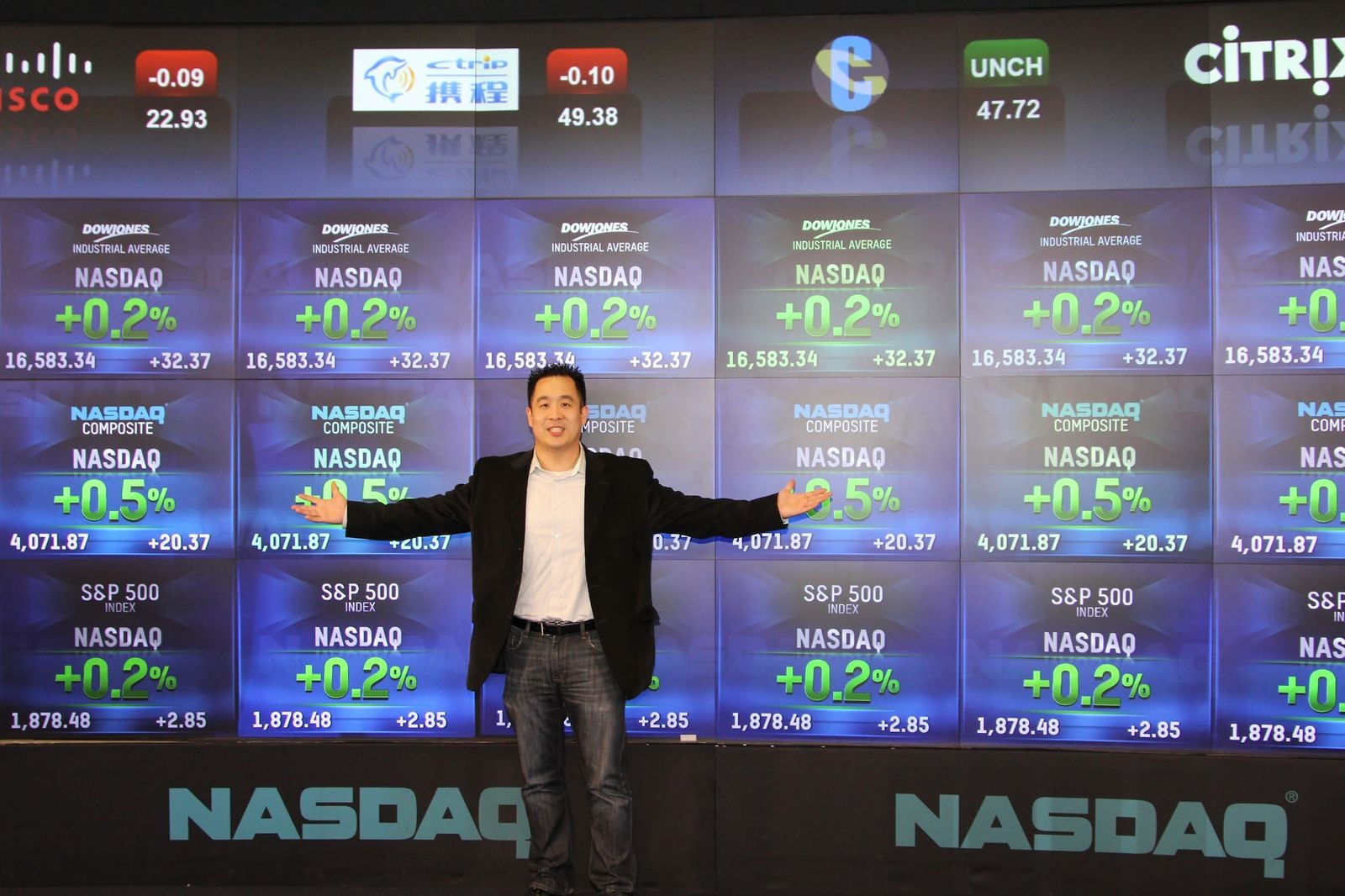 Tejune Kang at the Nasdaq MarketSite, arms out, beneath the live composite ticker board